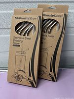 Two unopened packs of UltimateStraw stainless steel drinking straws, showing the front packaging with a cutout window revealing the straws inside.