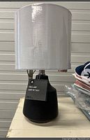 Photo of new black table lamp with white drum shade on a surface, showing tags attached and lamp in new condition.