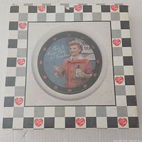 Photo of the I Love Lucy wall clock in original packaging showing box design with checkered black and white pattern and small red hearts with 'I Love Lucy' text. Window reveals clock face featuring Lucy holding a bottle and spoon.