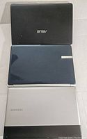 Three closed laptops stacked with Asus on top, unidentified dark laptop in middle, Samsung at bottom.