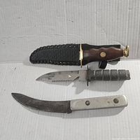 Three vintage knives arranged on white background. One wooden handle knife in leather sheath, one grey handle serrated blade knife, one white handle curved blade knife.