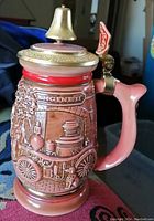 Stein showing the detailed front relief of a fire engine with a pink handle and brass lid with small bell.