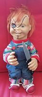 Front view of the Chucky doll with visible stitched facial scars, red hair, striped shirt, denim overalls, and plastic knife.