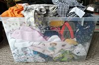 Plastic bin filled with various infant to toddler garments including printed onesies and patterned clothing.