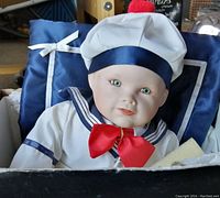 Front view of the Matthew doll dressed in a white sailor outfit with a navy blue ribbon on the hat and a red bow tie, seated on a blue satin pillow.