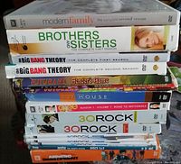 Stack of 10 DVD sets showing titles and seasons clearly including Modern Family, Brothers and Sisters, The Big Bang Theory (two seasons), Futurama, House, Glee, 30 Rock (two volumes), Dexter, and Arrested Development.