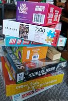 Photo showing stacked board games and puzzle boxes including FRIENDS 1000-piece puzzle, Live Laugh Lose, All In The Family, Storage Wars, Monopoly special edition