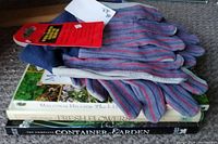 Photo showing three pairs of blue and gray work gloves with red stripes lying on top of three gardening books stacked on the floor. The glove packages have red tags.