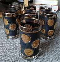 Front view of six of seven Libbey Glass Co. vintage cocktail glasses with black background and raised gold Old World coin motifs.