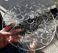 Clear glass plate held near window to highlight embossed grapes and leaf pattern on surface, showing the detail and clarity of glass.