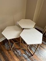 Set of three hexagon nesting tables with white tops and gold metal legs, arranged in the corner of a room on wood flooring.