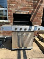 Front view of stainless steel BBQ grill with open lid showing cooking grates. Grill has four control knobs, side tables, and two-door storage cabinet below.