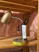 Desk lamp with gold lampshade and gooseneck attached to round metal LED base, black power cord visible.