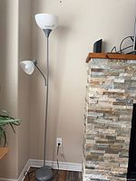 Full view of the silver metal floor lamp standing next to a wall and stone fireplace. Shows both the main upward-facing light with frosted glass bowl shade and the smaller adjustable reading light attached below with frosted glass shade.