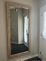 Full-length rectangular hallway mirror with a light wood-tone frame mounted on the wall beside a door and reflecting part of the hallway interior.