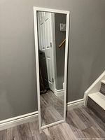 Full-length rectangular mirror with white frame placed against gray wall near stairs, clear reflective surface visible.