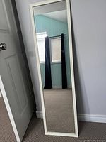 Full-length mirror with beige frame leaning against a wall, reflecting a room corner with a window and black curtains.