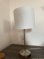 Full view of first night table lamp with white fabric shade and brushed metal base, pull chain visible