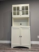 Photo of a white pantry storage unit with dual-door glass upper cabinets, open middle shelf with cable hole, and dual-door solid lower cabinets, in a room with gray walls and wood floor.