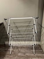 Image of a white foldable drying rack with three tiers of horizontal bars for clothes drying, standing on a wood floor against a plain wall.