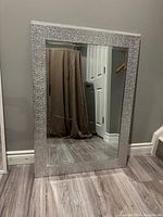 Rectangular mirror with a textured silver frame leaning against a wall, clear reflection visible