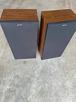 Pair of Sony speakers with dark wood finish and black front grille cloth placed on concrete floor.