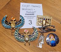 Top view of all 5 pieces arranged on a wooden surface with label reading "Egypt-Themed Brooches + Pendant"