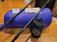 Photo showing two black Hammers trekking poles with foam grips placed next to a blue sleeping bag and black DeerFamy sleeping mat bag on wooden floor.