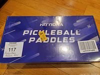 Blue rectangular unopened retail box labeled 'Pickleball Paddles' by Acttona with barcode and stickers on top, placed on wooden floor.
