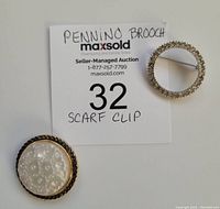 Both jewelry pieces placed on a white surface next to a MaxSold auction label identifying them as a Pennino brooch and scarf clip.