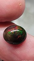 Close-up photo of a natural Ethiopian black opal held between fingers, showcasing vivid red, green, and orange play-of-color on a dark base.