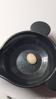 Oval natural Ethiopian opal gemstone placed in a black dish, showing visible flashes of color typical of high-quality opals, photographed under neutral lighting.