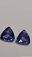 Close-up of two trillion cut tanzanite gemstones with a deep blue to purple coloration and high luster, shown on a neutral background from the front side.