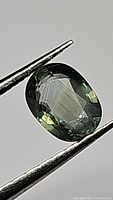 Close-up image of an oval cut moss green sapphire held by tweezers, showing the gemstone's facets and color