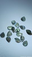 Overview of approximately fifteen natural pear cut alexandrite gemstones scattered on a white surface under daylight showing greenish hues.