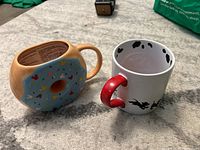 Two Disney mugs placed side by side: one shaped like a donut with Mickey Mouse theme, the other white ceramic with 101 Dalmatians puppy designs and red handle.