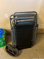 Folded black zero gravity lawn chairs stacked, one with detached black plastic cup holder accessory with two circular slots and one rectangular slot for attachment to chair armrest.