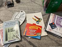 Art supplies on carpet including instructional drawing books, paint palette, Laurentian colored pencils, craft scissors, sketch pads, pastels, and watercolor paper