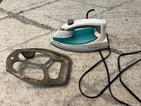 White and teal Black & Decker steam iron with attached cord and metal stand placed beside it on a floor surface.