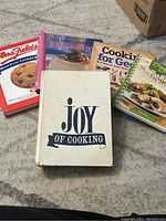 Five cookbooks shown together on the floor: Mrs. Fields Best Ever Cookie Book, The Best Ever Chocolate, Cooking for Greeks, Company's Coming Table for Two, and vintage Joy of Cooking with a cream cover.