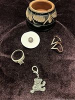 Photo of various items including a metal ring marked 925, triangle earrings, black and beige decorated container, silver-tone bear charm, and a small button or backing.