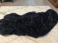 Black fuzzy blanket draped and spread on a light-colored rug showing size and texture.