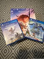 Front-facing photo of three Horizon PS4 game cases placed on a patterned fabric surface: Horizon Zero Dawn Complete Edition, Horizon Forbidden West, and a steelbook or art folder.