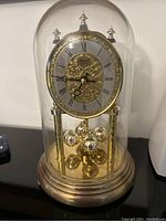 Front angled view of vintage Hermle clock under glass dome with ornate gold and grey dial, brass base and rotating pendulum.