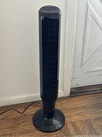 Front view of the black Climate Keep tower fan standing on a wooden floor against a wall near a door.