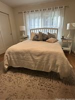 Photo showing queen bed with white wooden headboard and footboard, cream bedspread, multiple pillows, bedside tables, lamps, and area rug.