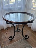 Front side view of the round table showing clear glass top, dark wood frame, and wrought iron curved legs with curl design, in a room with natural light.