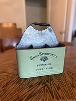 Front view of metal wine caddy showing front decorative text and grapes, green painted base and metal back panel with wood handle.