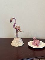 Photo of a tall pink glass flamingo on a white textured base and a small pink glass flamingo on a white dish on a dark wooden surface