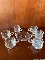 Photo showing six clear glass punch cups arranged on a round clear glass plate, all having circular beaded loop handles and a beaded edge on the plate.